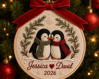 Puede incluir: Un adorno de bastidor de bordado redondo con dos pingüinos con bufandas verde y roja, enmarcados por ramas de pino y un copo de nieve bordados. Debajo de los pingüinos, el texto dice Jessica, un corazón, David y 2026. Un lazo de terciopelo rojo está sujeto en la parte superior.