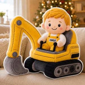 Op de afbeelding: Een zachtgele speelgoedgraafmachine met een pop in gele overalls, met de naam "Charlie" zichtbaar. Het speelgoed heeft een grijze bak en zwarte rupsbanden. De achtergrond is vervaagd met kerstverlichting.