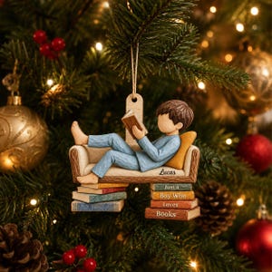 May include: A hanging Christmas ornament featuring a figure in blue pajamas reclining on a couch, reading a book. The couch is supported by stacks of books with the text "Lucas" and "Just a Boy Who Loves Books" printed on the side.