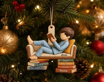 Könnte beinhalten: Ein hängender Weihnachtsbaumschmuck mit einer Figur in blauem Schlafanzug, die auf einem Sofa liegt und ein Buch liest. Das Sofa ruht auf Bücherstapeln mit dem Text "Lucas" und "Just a Boy Who Loves Books" an der Seite.
