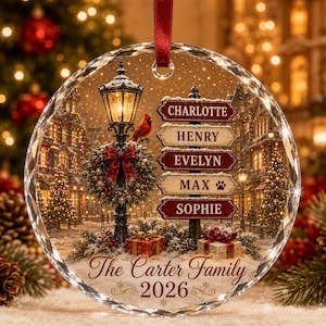 May include: A round, faceted glass Christmas ornament featuring a winter street scene with a lamppost, a red cardinal, and a signpost listing the names Charlotte, Henry, Evelyn, Max, and Sophie. It includes the text The Carter Family 2026.