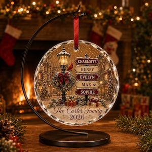 May include: A circular glass ornament hanging from a black metal stand, featuring a winter street scene with a lamppost, a red cardinal, and five stacked signs labeled Charlotte, Henry, Evelyn, Max with a paw print, and Sophie, plus The Carter Family 2026.