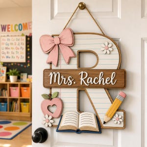 May include: A wooden door sign shaped like the letter R, featuring a lined paper pattern, a pink bow, a pink apple with a heart cutout, a yellow pencil, an open book, and white flowers. A brown horizontal plaque displays the text Mrs. Rachel.