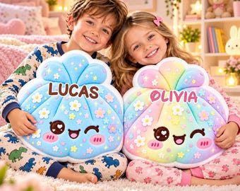 Puede incluir: Dos cojines de peluche en forma de concha con caras bordadas, mejillas sonrosadas y pequeñas flores aplicadas. Un cojín es azul claro con el nombre LUCAS y el otro es un degradado de arcoíris pastel con el nombre OLIVIA.
