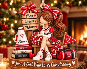 Puede incluir: Un adorno decorativo con una figura de animadora de cabello castaño y uniforme rojo y blanco, sentada junto a un letrero que dice Cheer, Dream, Believe. Incluye un megáfono con la etiqueta Emma, libros, pompones y una base con el texto Just A Girl Who Loves Cheerleading.