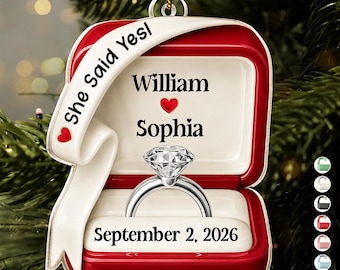 Puede incluir: Un adorno en forma de caja de anillo roja y blanca con un gráfico de anillo de diamantes, el texto "She Said Yes!", los nombres "William" y "Sophia" separados por un corazón rojo y la fecha "September 2, 2026" en la base.
