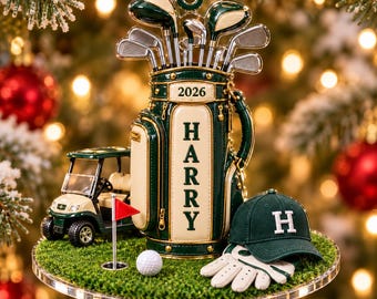 Puede incluir: Un adorno decorativo que presenta una bolsa de golf de color verde oscuro y crema con el nombre HARRY y el año 2026 impresos. El conjunto incluye un carrito de golf en miniatura, una pelota, una bandera y una gorra verde con la letra H.