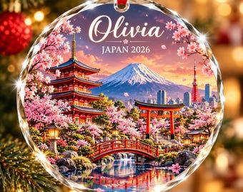 Puede incluir: Un adorno de cristal redondo y transparente con una escena impresa de una pagoda japonesa, un torii rojo, un puente sobre el agua y el monte Fuji. El texto "Olivia" y "JAPAN 2026" está impreso en la parte superior. Cuelga de una cinta roja.