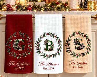 Puede incluir: Tres paños de cocina de tejido gofrado en rojo oscuro, blanco y beige claro. Cada uno presenta una corona navideña impresa que rodea una letra grande G, B o S, con el texto The Grahams, The Browns o The Smiths y Est. 2026.