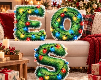 Puede incluir: Tres cojines de peluche con forma de letras E, O y S, cubiertos con una textura de agujas de pino verdes y envueltos con luces navideñas brillantes y coloridas en tonos rojo, azul, amarillo y verde en un entorno festivo.