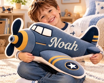 Puede incluir: Un avión de peluche en tonos azul marino, crema y amarillo. El lateral del avión presenta el nombre Noah en letras cursivas blancas. El diseño incluye una hélice, ventanas de cabina y un emblema circular con una estrella blanca.
