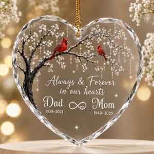 May include: A heart-shaped clear glass ornament featuring a printed tree with two red cardinals, white blossoms, and the text Always & Forever in our hearts, Dad 1938-2021, Mom 1940-2025, and an infinity symbol with a small heart in the center.