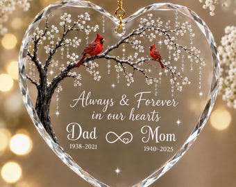 Puede incluir: Un adorno de cristal transparente en forma de corazón con un árbol impreso, dos cardenales rojos, flores blancas y el texto Always & Forever in our hearts, Dad 1938-2021, Mom 1940-2025, junto a un símbolo de infinito con un corazón.