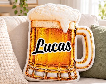 Puede incluir: Un cojín con forma de jarra de cerveza llena de líquido dorado y una espesa espuma blanca. El centro de la jarra presenta el nombre Lucas impreso en una fuente cursiva negra y negrita sobre el fondo de color ámbar.