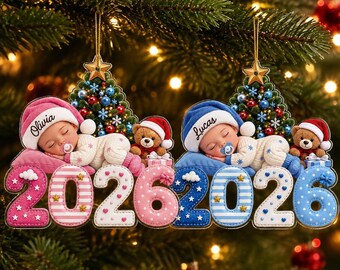 Puede incluir: Dos adornos navideños con bebés durmiendo con gorros que dicen Olivia y Lucas. Cada bebé descansa sobre una almohada junto a un osito de peluche y un árbol decorado. El año 2026 aparece en números grandes y estampados en la base.