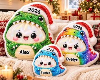 Puede incluir: Tres cojines de peluche con forma de albóndigas blancas que llevan disfraces de dinosaurio de colores y gorros de Papá Noel. Los cojines llevan el texto 2026 en los gorros y los nombres Alex, Evelyn y Sophia en las barrigas.