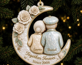 Puede incluir: Un adorno en forma de media luna con una pareja de ancianos sentados juntos. La luna está decorada con rosas blancas, perlas y el texto Emma, Daniel, Together Forever, Since 2004. La pareja viste una chaqueta crema y una gorra azul.