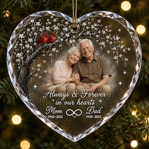 May include: A heart-shaped clear acrylic ornament featuring a printed image of an older couple, two red cardinals on a flowering tree, and the text Always & Forever in our hearts Mom 1940-2021 Dad 1938-2025 with an infinity symbol.