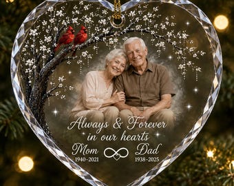 Puede incluir: Un adorno de acrílico transparente en forma de corazón con la imagen impresa de una pareja mayor, dos cardenales rojos en un árbol en flor y el texto Always & Forever in our hearts Mom 1940-2021 Dad 1938-2025 con un símbolo de infinito.