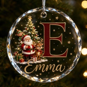 May include: A circular glass Christmas ornament with a faceted edge, featuring a large red letter E, the name Emma in gold script, a decorated Christmas tree, Santa Claus, a reindeer, and an elf, all surrounded by small gold stars.