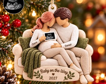 Puede incluir: Un adorno colgante con una figura tallada de una pareja sentada en un sofá. La mujer tiene el pelo rosa y el hombre el pelo castaño. Sostienen una pequeña ecografía. La base del sofá está grabada con Baby on the Way 2027.
