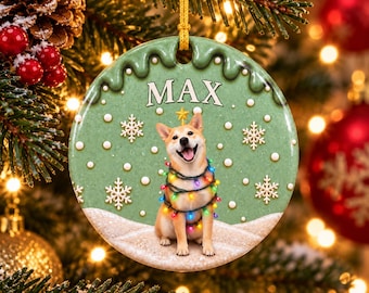 Puede incluir: Un adorno navideño redondo de cerámica con fondo verde que muestra a un perro Shiba Inu envuelto en luces de colores. El nombre MAX está impreso en blanco en la parte superior. El diseño incluye copos de nieve blancos y una base nevada.