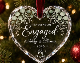 Puede incluir: Un adorno de cristal transparente en forma de corazón con bordes facetados, colgado de una cinta de terciopelo rojo oscuro. Presenta hojas de eucalipto verdes, el texto THE YEAR WE GOT Engaged, Ashley & Thomas, 2026 y dos copas de champán.
