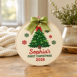 May include: A circular embroidery hoop ornament featuring a stitched green Christmas tree with yellow ornaments, red and green snowflakes, and a green satin bow. The text reads Sophia's FIRST CHPISTMAS 2026 in red and green embroidery thread.