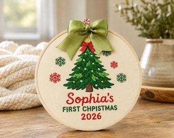 Puede incluir: Un adorno en bastidor de bordado circular con un árbol de Navidad verde cosido, adornos amarillos, copos de nieve rojos y verdes, y un lazo de satén verde. El texto dice Sophia's FIRST CHPISTMAS 2026 en hilo de bordar rojo y verde.