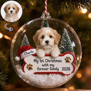 May include: A clear spherical Christmas ornament containing a light-colored puppy, two miniature green pine trees, and artificial snow. A bone-shaped plaque reads "My 1st Christmas with my forever family 2026" with "Charlie" on a red Santa hat.