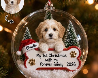 Puede incluir: Un adorno navideño esférico transparente que contiene un cachorro de color claro, dos pinos verdes en miniatura y nieve artificial. Una placa en forma de hueso dice "My 1st Christmas with my forever family 2026" y "Charlie" en un gorro de Papá Noel.
