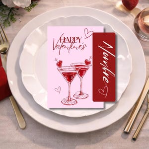 May include: A Valentine's Day card and bookmark on a white plate setting. The pink card says "Happy Valentine's" with martini glasses and hearts. The red bookmark has a name written on it. Gold cutlery is on the side.