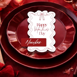 Puede incluir: Una romántica mesa de San Valentín con platos burdeos. Una tarjeta blanca con un lazo y las palabras "Happy Valentine's Day" y "Nombre" se coloca sobre los platos. Velas y pétalos de rosa añaden ambiente.