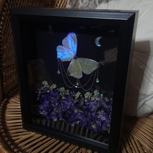 Puede incluir: Obra de arte enmarcada con una mariposa de alas azules iridiscentes, adornada con una cadena plateada y detalles de lágrima. Sobre un fondo oscuro con flores moradas y una luna creciente, en un marco negro.