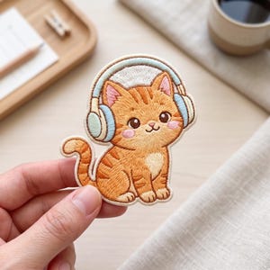 May include: An embroidered patch featuring an orange tabby cat with dark stripes, wearing light blue and white over-ear headphones. The cat has large dark eyes, pink cheeks, and a small white patch on its chest, rendered in detailed thread work.