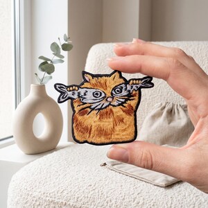 May include: An embroidered patch featuring a fluffy orange cat holding a silver fish horizontally across its face. The cat has detailed orange fur stitching, pointed ears, and a black border. The fish is grey with black outlines and visible scales.