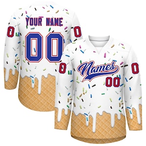 Puede incluir: Jersey de hockey blanco con un diseño de cono de helado. El jersey presenta una base de gofre, glaseado blanco derritiéndose y chispas de colores. La parte posterior muestra el texto "YOUR NAME" sobre el número "00" en azul. La parte delantera tiene la palabra "Names" en azul.