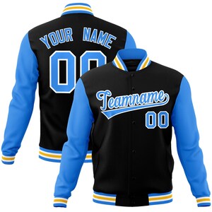 May include: Black varsity jacket with light blue sleeves. The jacket features the text "Your Name" and "Teamname" in white script, along with the number "00". The collar and cuffs have yellow and white stripes.