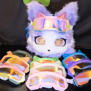 May include: A plush, lavender-colored fox head with cat-ear goggles. Several pairs of colorful, cat-ear-shaped goggles in shades of pink, orange, green, and blue are arranged in front of the fox head.