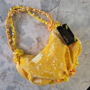 May include: A yellow handbag with a floral pattern, featuring a braided handle adorned with pearls and gold beads. The bag has a ruffled trim and a tag attached.