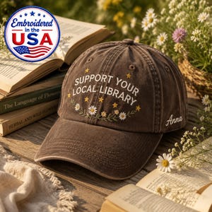 May include: A brown baseball cap featuring the embroidered text SUPPORT YOUR LOCAL LIBRARY in white, accented with yellow stars and white daisy flowers. The name Anna is embroidered in white on the side of the cap, which has a curved brim.