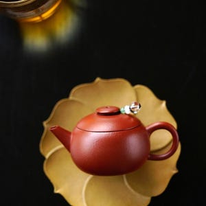 May include: A small, round, reddish-brown clay teapot with a matching lid sits centered on a golden-yellow, petal-shaped saucer. A decorative bead charm with a clear crystal and green accent is attached to the lid handle of the teapot.