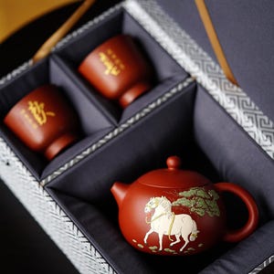 May include: A red clay teapot featuring a white horse and green tree illustration, accompanied by two matching red clay cups with gold Chinese characters, all housed in a gray fabric-lined gift box with a patterned exterior.