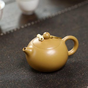 May include: A round, matte ochre-colored teapot with a smooth finish, featuring a small, curved spout, a circular handle, and a lid topped with a spherical knob and a small, white, flower-shaped decorative accent attached by a thin cord.