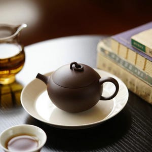 May include: A dark brown, matte-finish clay teapot with a rounded body, a curved spout, and a small loop handle on the lid. The teapot rests on a plain, off-white circular saucer, accompanied by a small white tea cup and a clear glass pitcher.