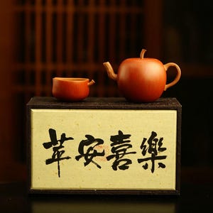 May include: A reddish-brown clay teapot shaped like an apple and a matching small cup resting on a rectangular box. The box features a light cream-colored front panel with black Chinese calligraphy characters printed across the center surface.