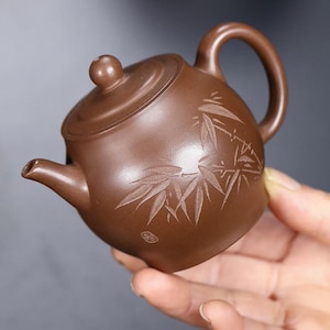 May include: A small brown clay teapot featuring a rounded body, a curved handle, a short spout, and a lid with a spherical knob. The exterior surface is decorated with an engraved bamboo leaf pattern and a small square seal mark near the base.