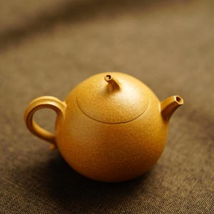 May include: A round, golden-yellow teapot with a textured, matte finish. It features a curved handle on the left, a short, angled spout on the right, and a small, pointed knob on the center of the lid. The body is spherical and uniform in color.