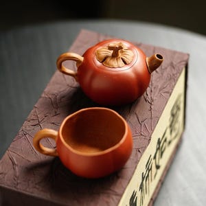 Puede incluir: Una tetera y una taza de arcilla de color marrón rojizo con una forma redondeada similar a una calabaza. La tapa de la tetera presenta una hoja y un tallo esculpidos. Ambos artículos descansan sobre una caja rectangular marrón texturizada.
