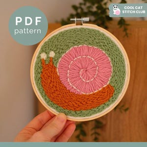 May include: Embroidered snail design in a wooden hoop. The snail features a pink spiral shell, orange body, and white antennae, set against a green background. The text "PDF pattern" is in a blue circle. The text "COOL CAT STITCH CLUB" is in the upper right corner.